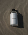 500ml navulfles magnesiumolie liggend op het strand, sfeervol gepresenteerd voor een gevoel van ontspanning en wellness. Verkrijgbaar in onze webshop voor dagelijks gebruik en eenvoudig navullen.