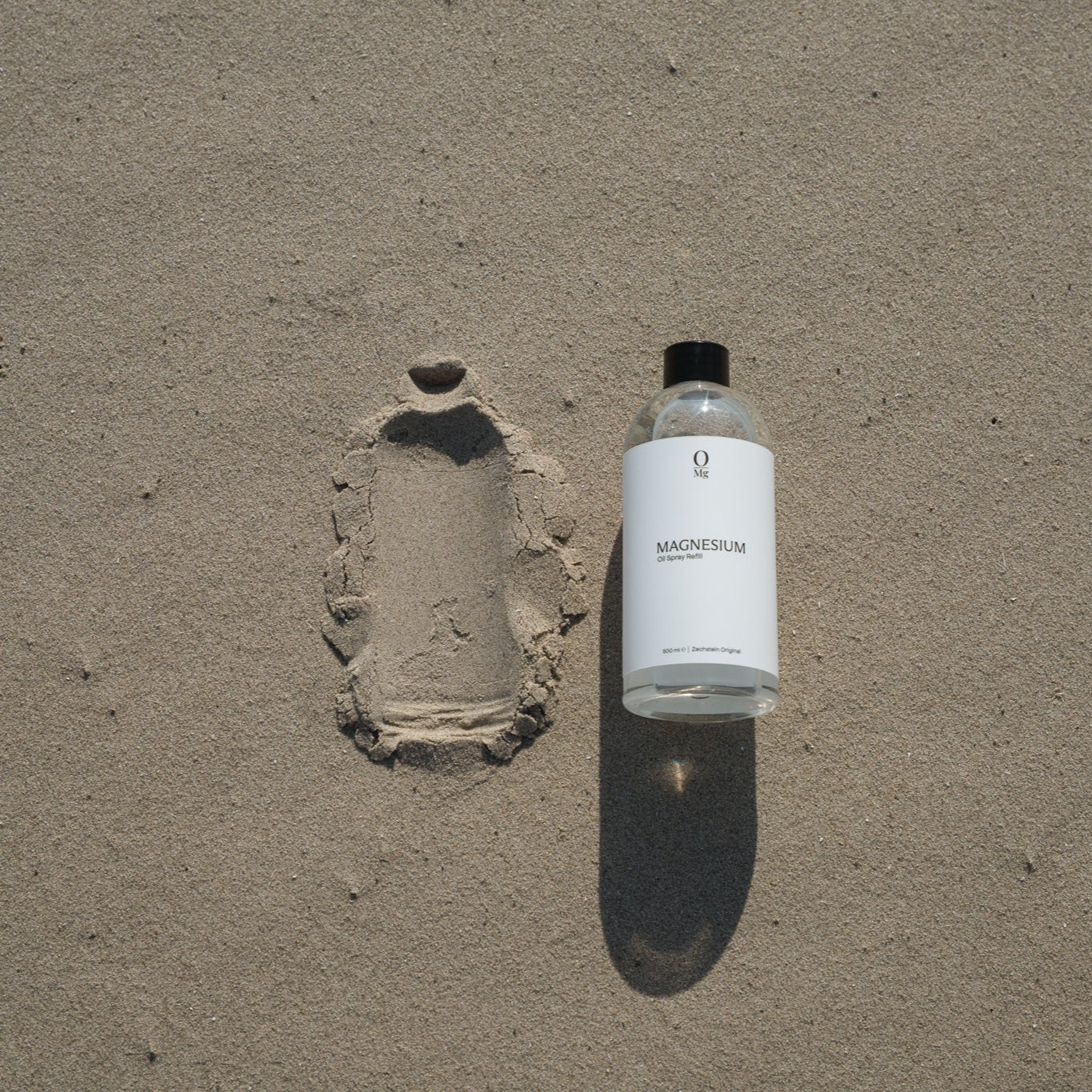 Foto van een 500ml navulfles magnesiumolie die op het strand ligt. Deze magnesium olie is goed voor ontspanning, huidverzorging en spierherstel, en eenvoudig te bestellen via onze webshop.